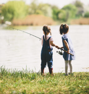 two little girls with a fishing pole on the bank of a lake 