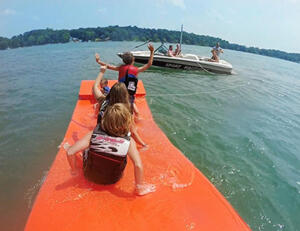 three children being pulled behind a boat on an orange watermat