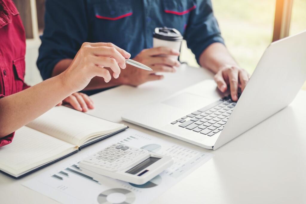 Two people looking at computer with calculator on desk and printed out spreadsheet