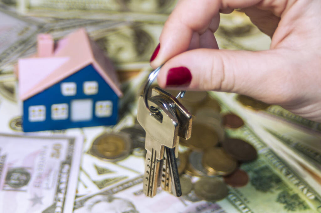 Woman holding set of keys over wooden house figurine and money