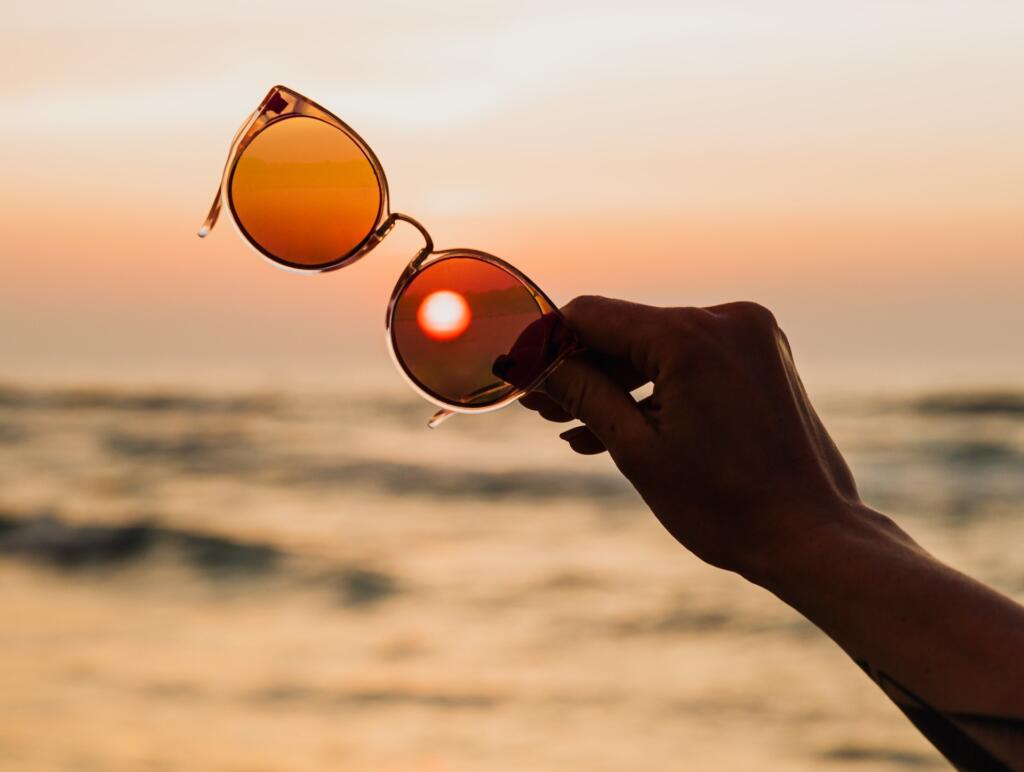 person holding sunglasses at sunset with the sun behind one lens