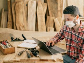 man in a workshop with wood, tools, a book, and a laptop