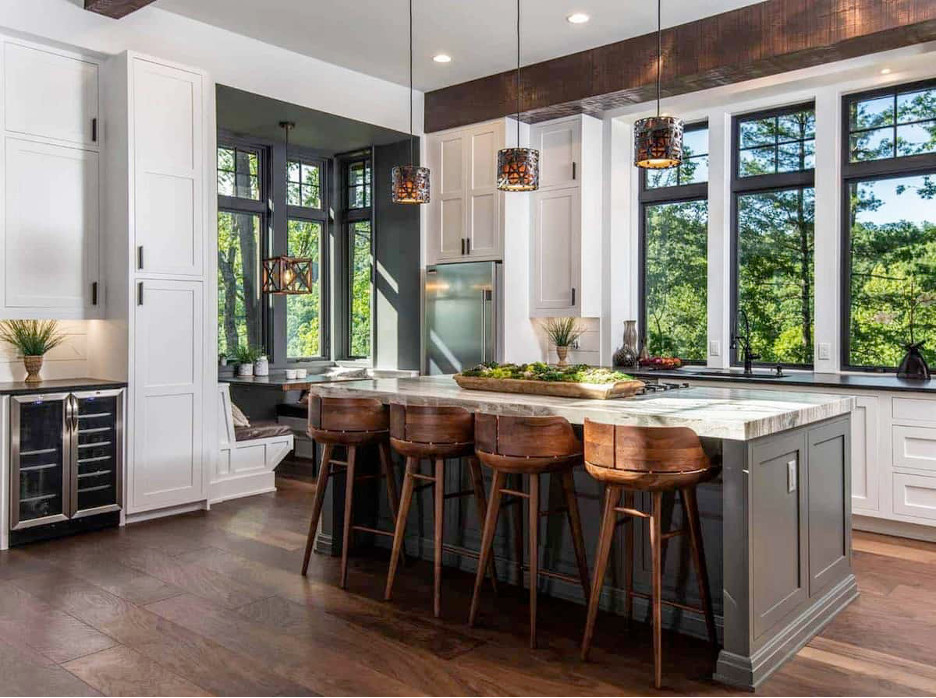 A cozy, rustic lake home kitchen with reclaimed wood floors, beams, and natural wood counter stools around a rustic stone island with view to outdoors in backdrop