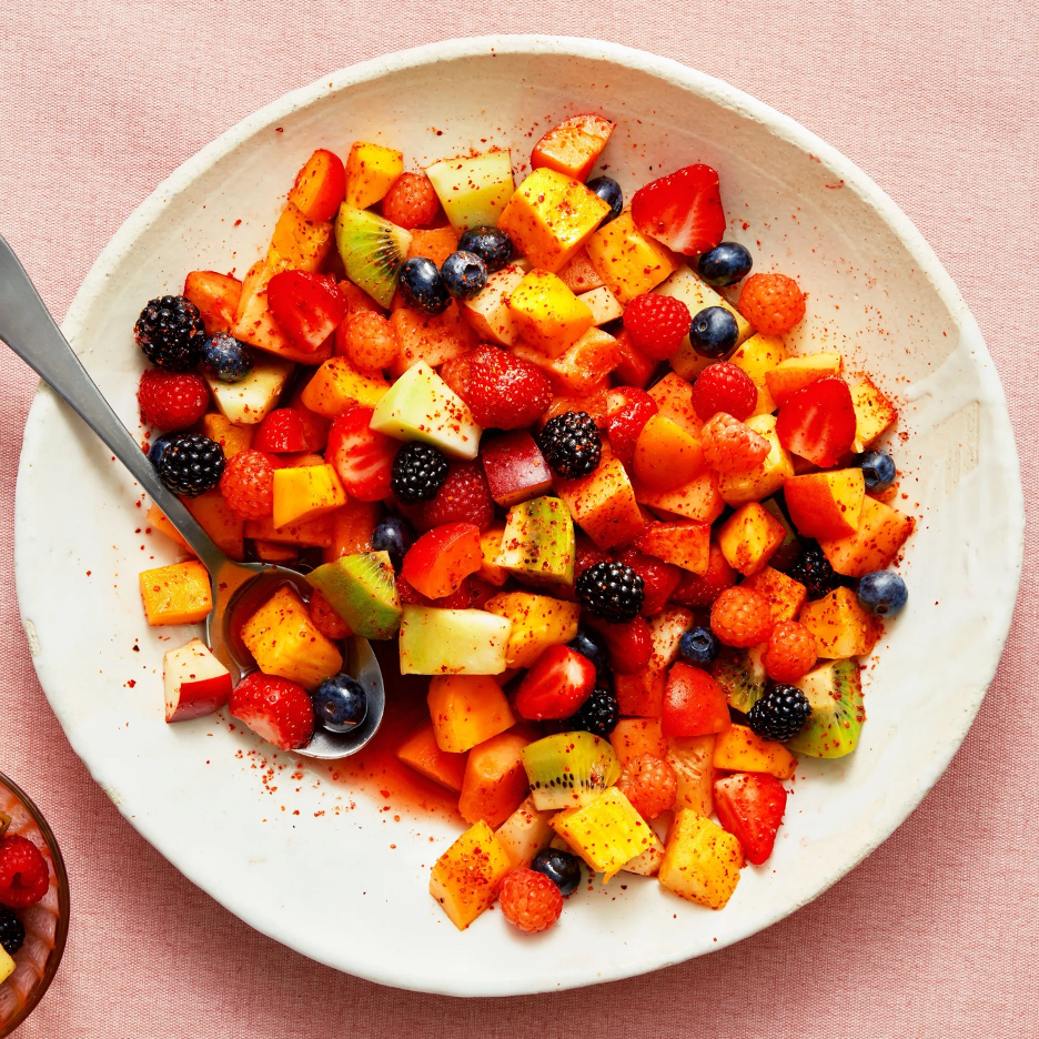 Honey and Bitters Fruit Salad in a white serving bowl with a spoon. Salad includes pineappele, strawberries, raspberries, kiwi, mango, and apples sprinkled with chili lime seasoning