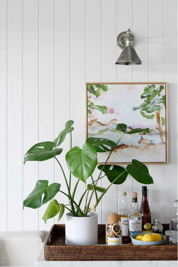 Pothos plant in white container set in a wicker tray holding bar essentials and a watercolor painting above