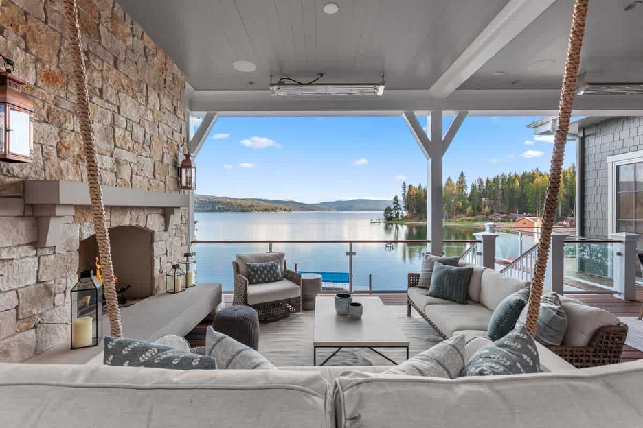 Multi-seasonal outdoor space at lake home with gorgeous lake view, stone fireplace wall, ceiling heating elements, neutral paette, swinging daybed, cushioned furnishings